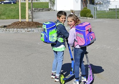 Zwei Kinder mit Ranzen auf dem Rücken und Tretrollern auf dem Schulweg
